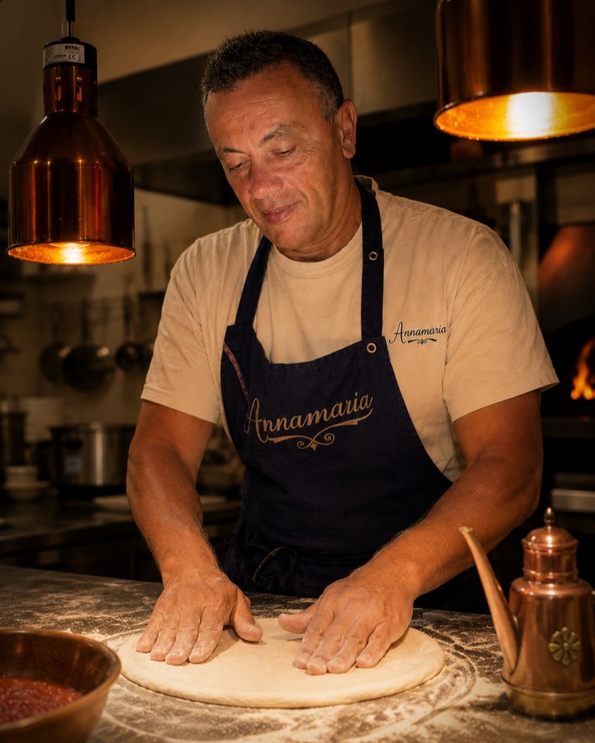 En cuisine : la pâte à pizza est étalée à la main sur le marbre fariné, sous les lampes chauffantes en cuivre, devant le four à bois